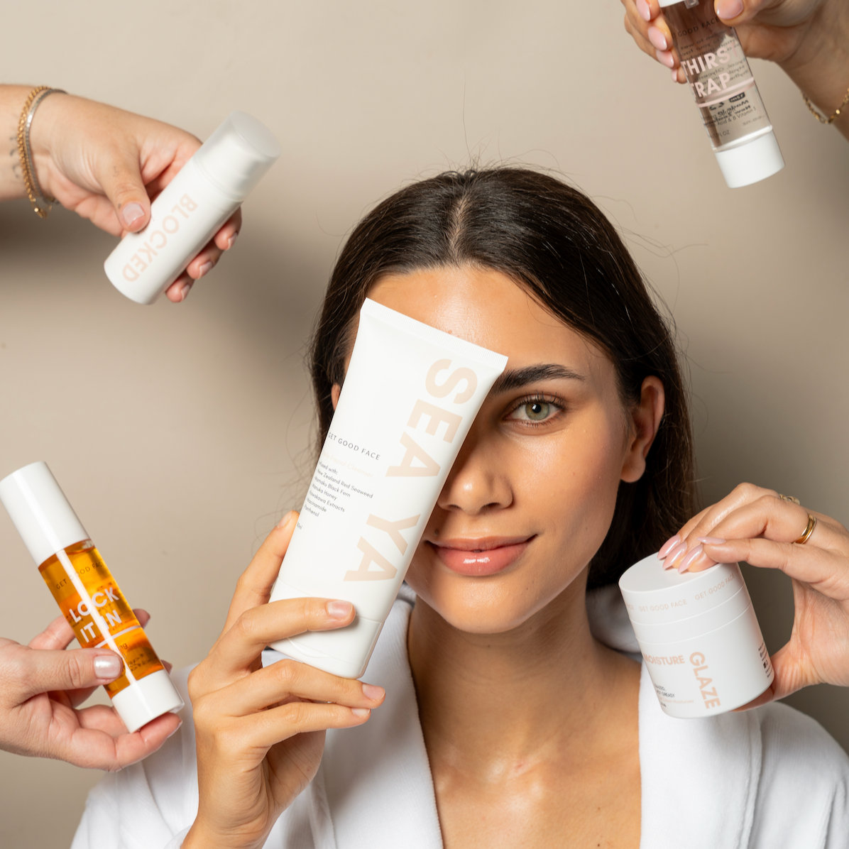 Woman in a white robe surrounded by skincare products against a beige background