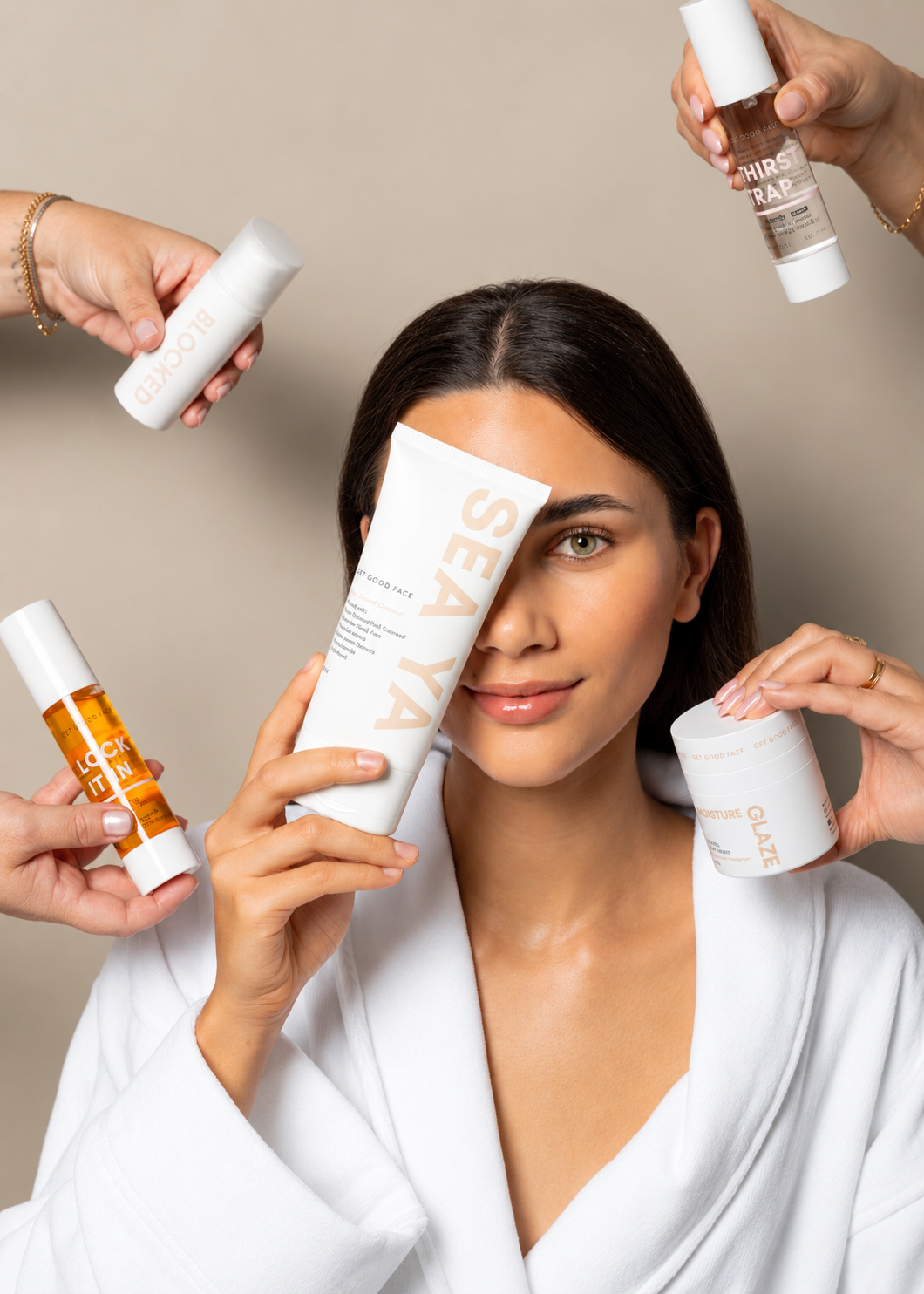 A woman in a white robe holds a large white tube labeled SEA YA in front of her face, surrounded by multiple hands holding additional SEA YA skincare products against a plain beige studio background.