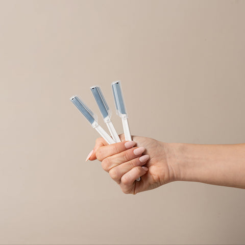 A hand holding three slim white tools with light blue tips against a plain beige background.