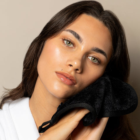 A woman with long dark brown hair and green eyes, wearing a white robe, holds a black textured facial cleansing cloth against her cheek against a neutral beige background.
