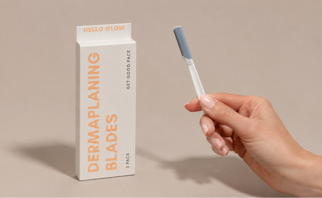 A hand holds a derma planing blade above a matching product box labeled 'DERMAPLANING BLADES' and 'HELLO GLOW' on a warm beige background.