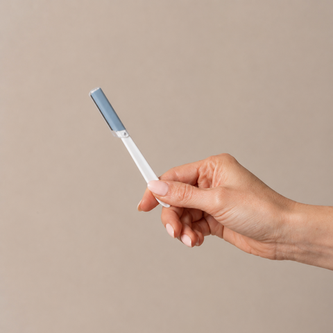A hand holding a slim white and light blue pen-like cosmetic tool against a plain warm beige background.